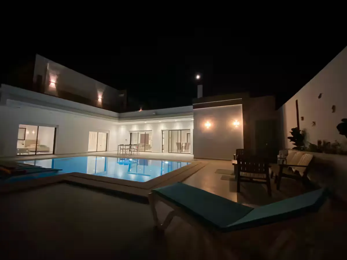 The villa at night: a lit private pool, sun loungers and a terrace in front of a white single-storey wing with glass doors.