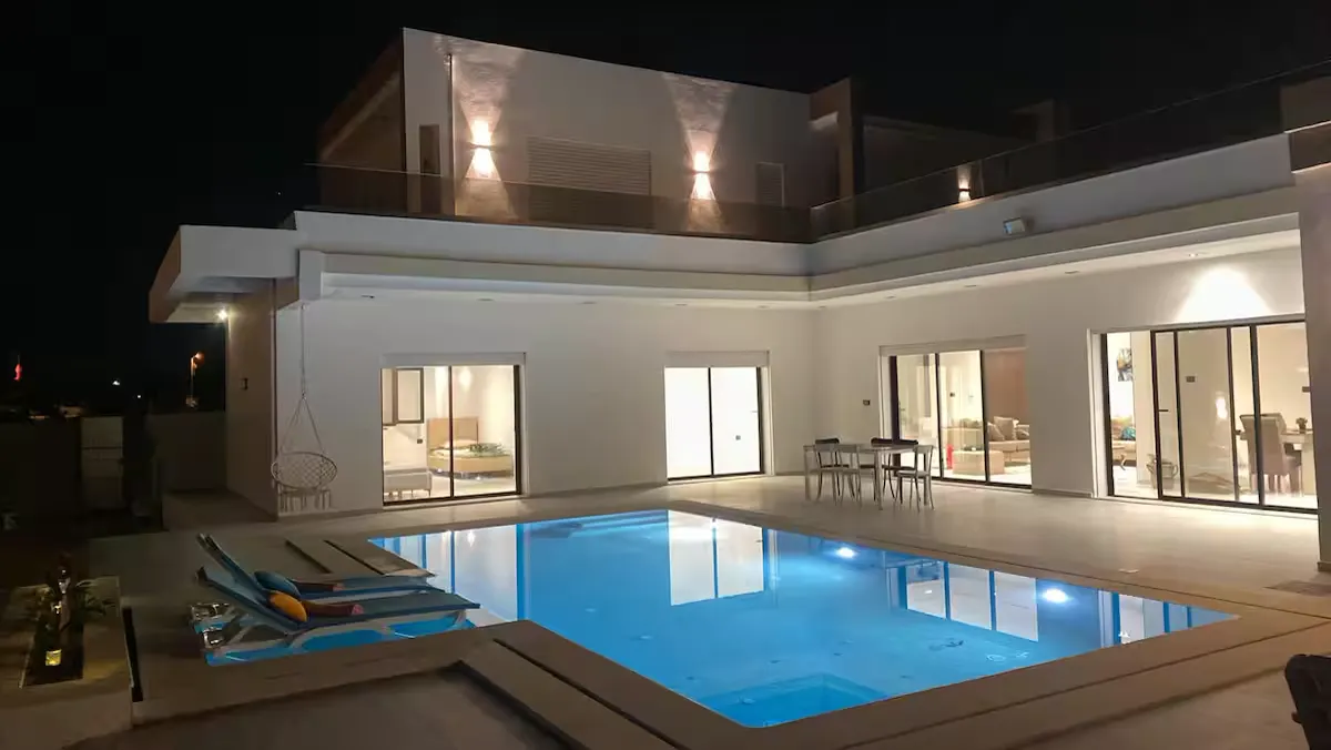 The two-storey white villa at night, its glass doors lit behind a private pool, a dining table and loungers on the terrace.